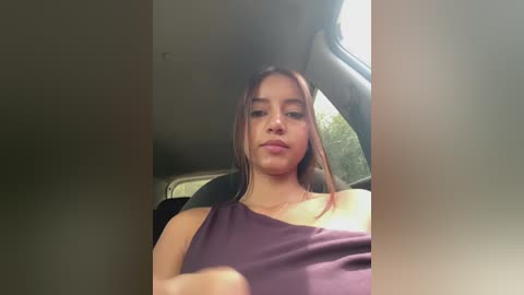 A video of a young woman with long, straight brown hair, wearing a sleeveless purple top, sitting in a car with a sunlit window. She has a neutral expression.