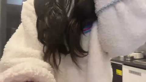 A close-up video of a person with long, wavy, dark hair in a white bathrobe, revealing a striking blue eye with purple and pink eyeshadow. The background shows a kitchen with a dark countertop and white appliances.