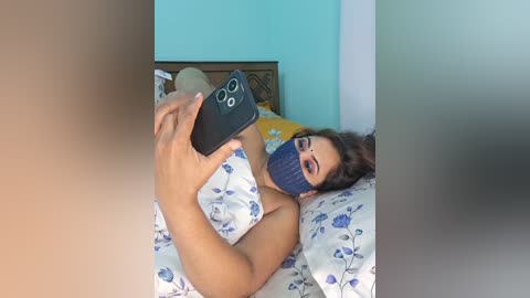 A video of a woman lying on a bed, wearing a blue face mask, blue floral-patterned pillowcase, and holding a black smartphone, capturing her reflection. The background features a turquoise wall and a wooden headboard.