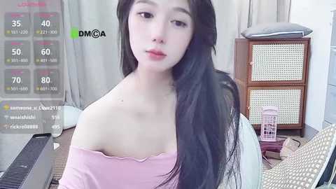 Video of a young Asian woman with long black hair and fair skin, wearing a pink off-shoulder top, sitting indoors beside a wooden cabinet.