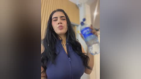 Video of a young woman with long black hair, wearing a sleeveless navy blue top, drinking from a blue water bottle, in a modern, brightly lit room with beige walls and a glass door.