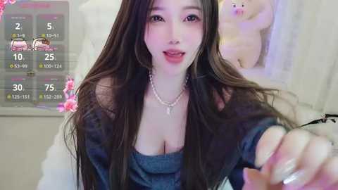 A video of an East Asian woman with long, dark hair and pale skin, wearing a low-cut blue dress, in a bedroom setting.