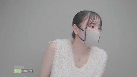 Video of an East Asian woman with straight black hair, wearing a white, fuzzy-textured sleeveless top, and a white surgical mask, set against a plain, light gray background.