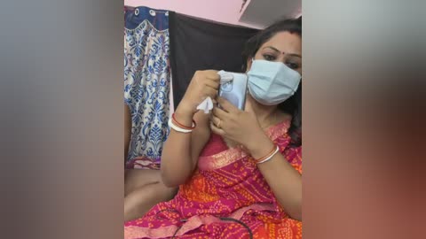 Video of a South Asian woman with medium skin tone, wearing a pink and orange saree, a blue face mask, and gold bangles. She sits on a bed, holding a blue tissue. Background includes a patterned curtain and a dark wall.