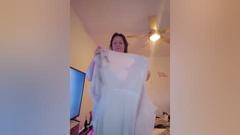 A video captures a woman with medium-length brown hair, wearing a white dress, standing in a dimly lit room with beige walls and a ceiling fan.