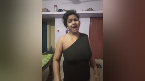 Video of an overweight woman with dark hair in a black one-shoulder dress, mouth open in a loud yell, in a cluttered bedroom with pink walls and a green bed.