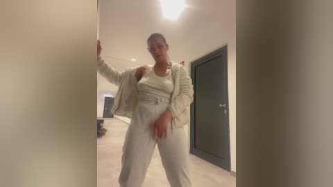 Video of a slim, dark-skinned woman with short hair, wearing glasses, a white sweater, and loose pants, standing in a modern hallway with beige walls and a black door.