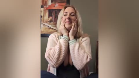 A video of a blonde woman with fair skin, wearing a light pink knitted sweater, resting her hands on her cheeks, smiling. Behind her, a painting of a rustic house in a French village style.