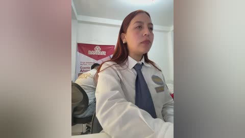 Video of a Latina woman with long, straight, auburn hair, wearing a white shirt and blue tie, standing in an office with a red and white banner in the background.