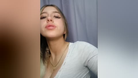 Video of a young woman with fair skin, light brown hair, wearing a light grey V-neck top and a gold necklace, puckering her lips in a kiss. Background is a plain, light-colored curtain.