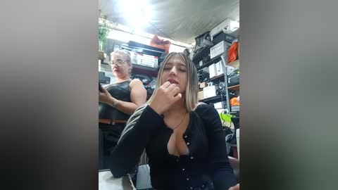 Video of two women in a cluttered storage room; one with blonde hair and a black top, biting her finger, while the other, with brown hair, stands behind a desk.