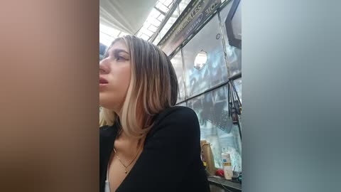 Video of a blonde woman with shoulder-length hair, wearing a black top, standing in a dimly lit, cluttered room with a window and various objects in the background.