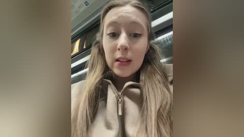 A video of a young, light-skinned woman with long, blonde hair in pigtails, wearing a beige jacket. She has a neutral expression. The background shows a subway car with fluorescent lighting.