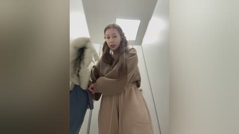 Video of a slender, fair-skinned woman with long, straight brown hair, wearing a beige coat, standing in a narrow, white-walled hallway with a white ceiling light, looking startled as she grabs the sleeve of a partially visible person.