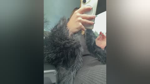 A video captures a woman in a black fur coat and gray sweater, seated in a car, holding a smartphone with her right hand.