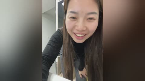 Video of a smiling East Asian woman with long black hair, wearing a black top, standing in a modern bathroom with a white sink and brown door.