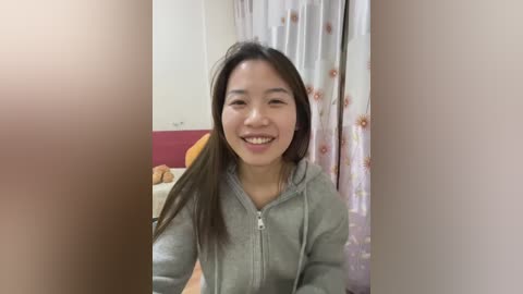 A video of a young Asian woman with long, straight black hair, smiling, wearing a light gray hoodie, standing in a bedroom with a pink curtain and a stuffed animal on a bed.
