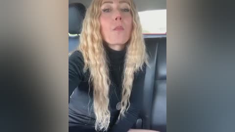 A video shows a blonde woman with wavy hair, wearing a black top, sitting in a car with a blurred background.