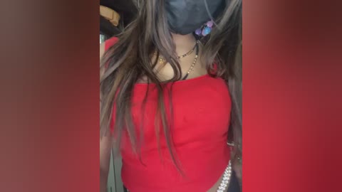 Media: Video of a person wearing a red long-sleeve top, black face mask, and a silver chain necklace. Long brown hair partially covers the face. Background is blurred in red.