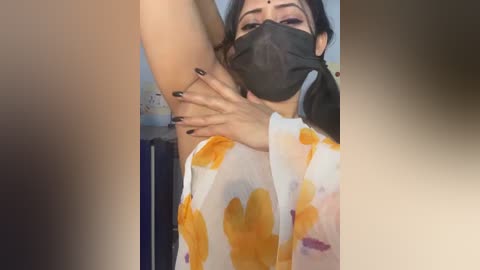 Video of a woman in a mask, with dark hair, wearing a white top with yellow and orange splatters, sitting on a blue chair, hands up, black nails, and blurred background.