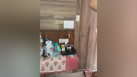 Video of a cluttered room with a white table covered in various items including a water bottle, a blue cup, a black bag, and a pink blanket. The wooden walls and ceiling contribute to a rustic feel.