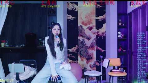 Video of an Asian woman with long black hair, sitting on a stool, singing into a microphone in a dimly lit room with a large, colorful mural behind her.