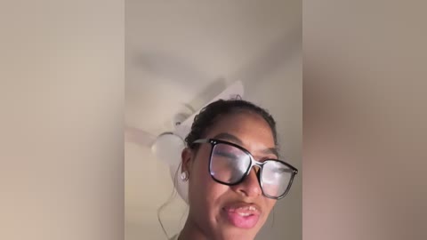 Video of a young Black woman with medium brown skin, wearing black-rimmed glasses and a white headband, smiling. Background is a plain, beige wall.