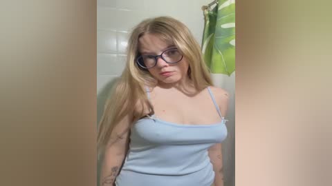 Video of a blonde woman with glasses, wearing a light blue spaghetti-strap top, standing in a tiled bathroom with a green shower curtain in the background.