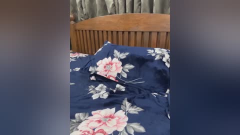 Video of a neatly made bed with a navy blue duvet cover adorned with large pink and white floral patterns. The wooden headboard and green curtains in the background add a cozy, homely feel.