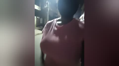 Video of a woman with dark skin wearing a pink shirt, partially obscured by shadows, in a dimly lit room with indistinct background objects.