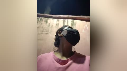 Video of a person with long dark hair, wearing a black face mask and a pink shirt, standing against a peeling, beige wall with a wooden beam overhead.