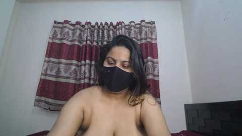 Video of a topless woman with medium brown skin and long black hair, wearing a black face mask, sitting on a bed with red and white striped curtains in the background.