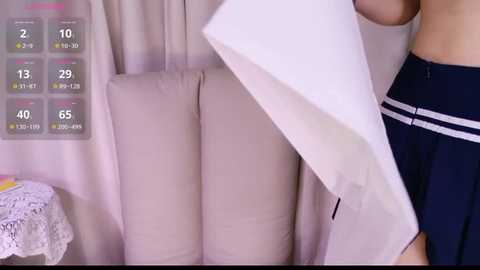 A video of a person in a dark navy skirt and white top, holding a white sheet, standing next to a beige couch with a digital thermometer display on the left.