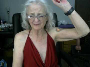 Media: Video of an elderly woman with white hair, glasses, and a red halter top, sitting on a black office chair. Background includes a desk with various items and a person partially visible.