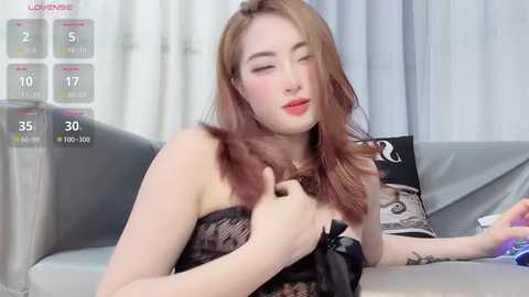 Video of a young, fair-skinned woman with long, wavy, light brown hair, wearing a black strapless lace dress, sitting on a gray couch. She has red lipstick and is touching her hair.