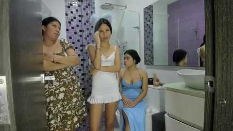 Video of three Asian women in a modern bathroom; one in a white dress, another in a blue dress, and a third in a floral dress, with tiled walls and a white sink.