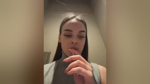 Video of a young woman with light skin and straight, long brown hair, wearing a gray turtleneck sweater, looking contemplative while holding a pen to her lips, indoors.