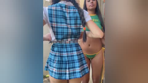 Video of two young women in a hallway: one wearing a blue plaid dress, the other a green crop top and yellow bikini bottoms.