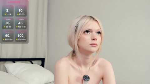 A video of a topless, fair-skinned woman with blonde hair, wearing a pendant necklace, sitting on a bed in a minimalistic, beige room.