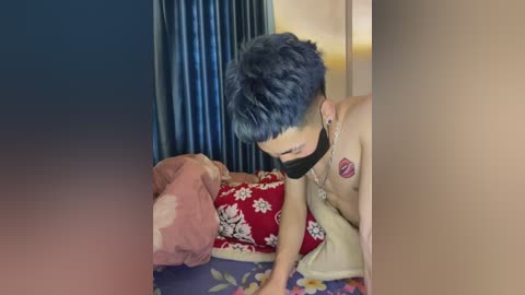 A video of a young Asian man with short blue hair, wearing a black mask and no shirt, leaning over a bed with red floral sheets, in a dimly lit room with a blue curtain.