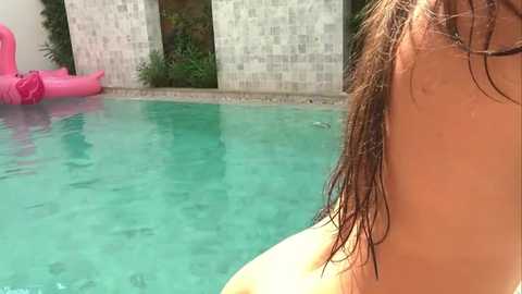 Video of a woman with wet, shoulder-length brown hair, partially submerged in a turquoise pool, facing away. In the background, a pink inflatable flamingo floats near beige brick walls.