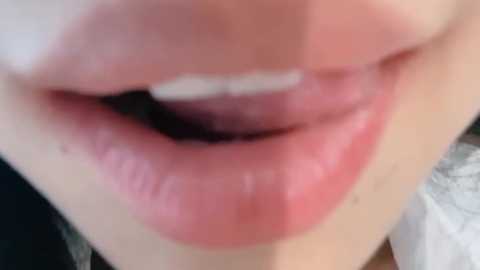 Close-up video of a person's lips, showing a slightly open mouth revealing teeth. The lips are glossy and slightly parted, with a natural pink hue. The background is blurred, with a hint of white fabric visible.