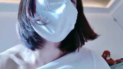 A video of a person with short, dark hair wearing a white surgical mask and white lab coat, standing indoors near a toy monkey on a shelf.