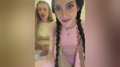 Video of two young women: a blonde with long hair and a brunette with braided hair, both wearing pastel outfits, in a dimly lit room.