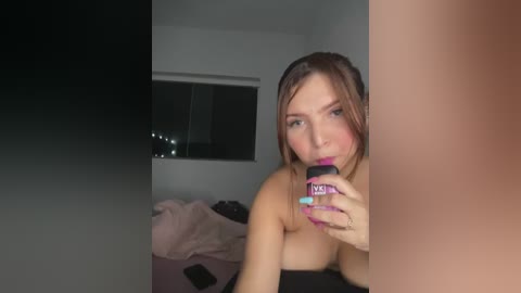 A video of a topless woman with fair skin, brown hair, and purple-painted nails, drinking from a purple bottle in a dimly lit bedroom.