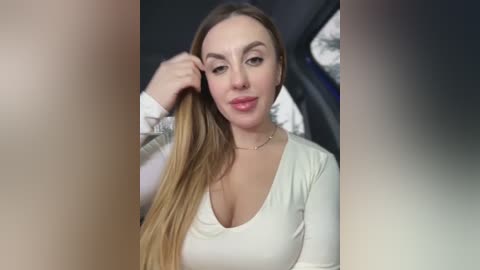 Video of a Caucasian woman with long blonde hair, fair skin, and brown eyes, wearing a white long-sleeve top, holding her hair, in a blurred background.