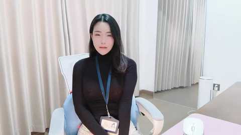 Video of an Asian woman with long black hair, wearing a black turtleneck, seated in a white chair in a room with beige curtains and light-colored walls.