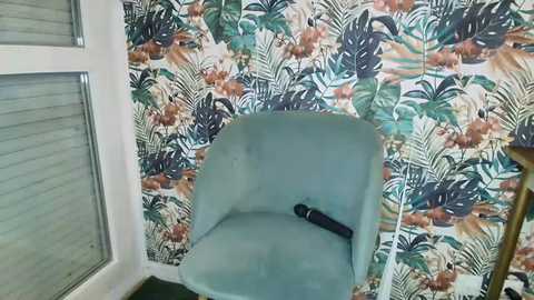 Video of a teal mid-century modern chair with a black umbrella, set against a vibrant, tropical-patterned wallpaper. The scene includes a window with horizontal blinds on the left.