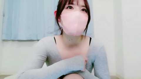 Video of an East Asian woman with short brown hair, wearing a light gray top, holding a pink mask to her mouth, in a room with white walls and blue curtains.