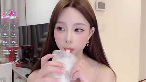 Media: Video of a young East Asian woman with long brown hair, wearing a pink spaghetti strap top, drinking from a plastic bottle. Background includes a kitchen countertop and modern decor.